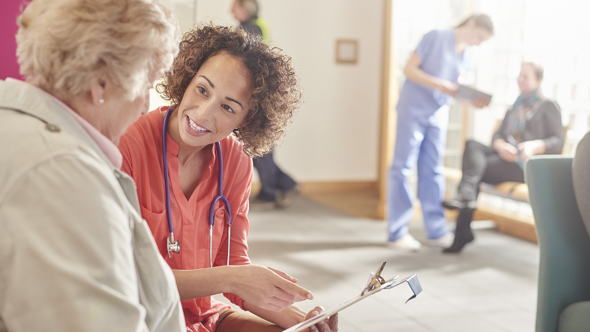 A female doctor stands next to a senior patient in her waiting room and casually chats to her about her details. In the background a nurse with a tablet and a patient at the desk and booking an appointment. A female doctor stands next to a senior patient in her waiting room and casually chats to her about her details. In the background a nurse with a tablet and a patient at the desk and booking an appointment.