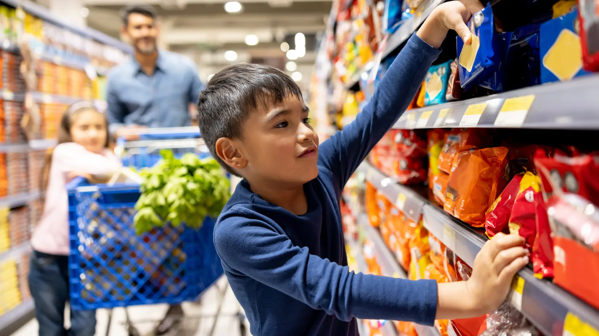 A young boy picking items from a shelf at the grocery store, with his family in the background.