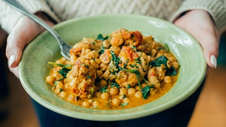 Hands holding a bowl of chickpea curry topped with fresh herbs.