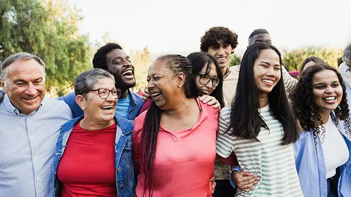A large group of diverse people stand outdoors while smiling and holding each other.