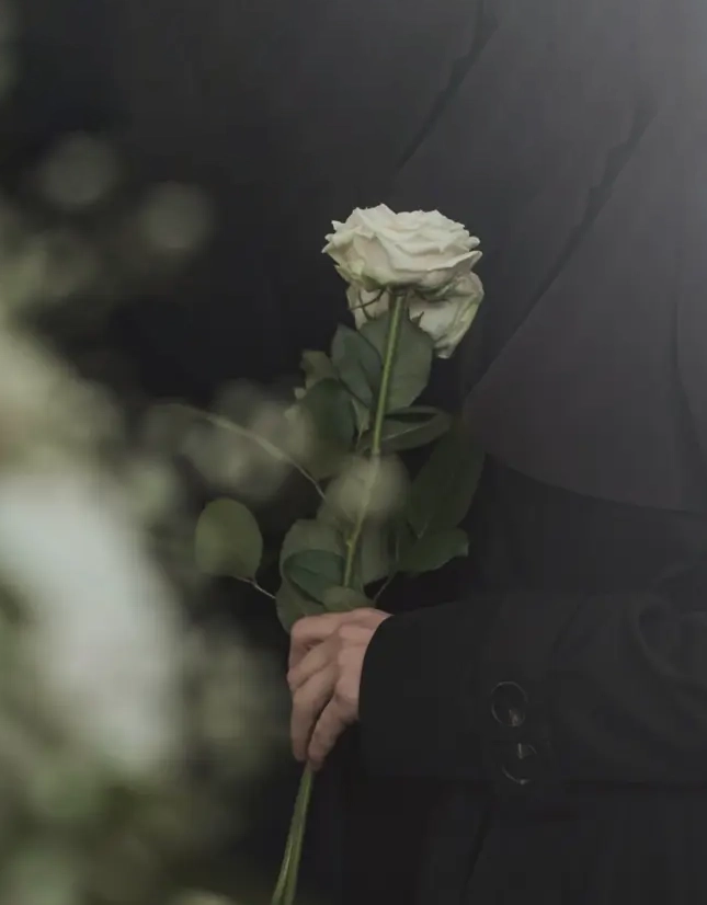 Person holding a single white rose at an outdoor memorial.​​​​