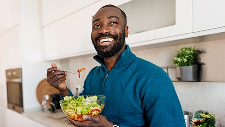 A person in a kitchen holding a bowl of salad and eating with a fork. 