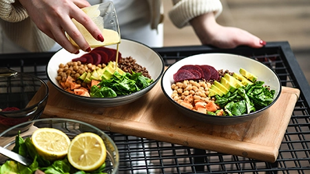 A person pours dressing onto two bowls filled with vegetables, grains and chickpeas. 