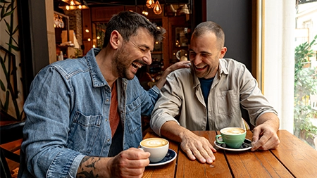 Two people sit together at a cafe table, talking over cups of coffee. 
