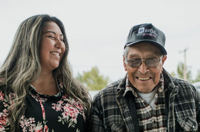 A younger woman smiles at an elderly man wearing a baseball cap.