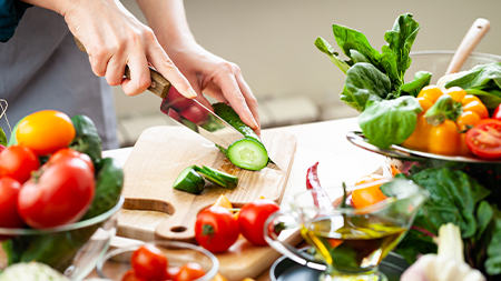 Person slicing a cucumber on a wooden, cutting board surrounded by fresh vegetables.
