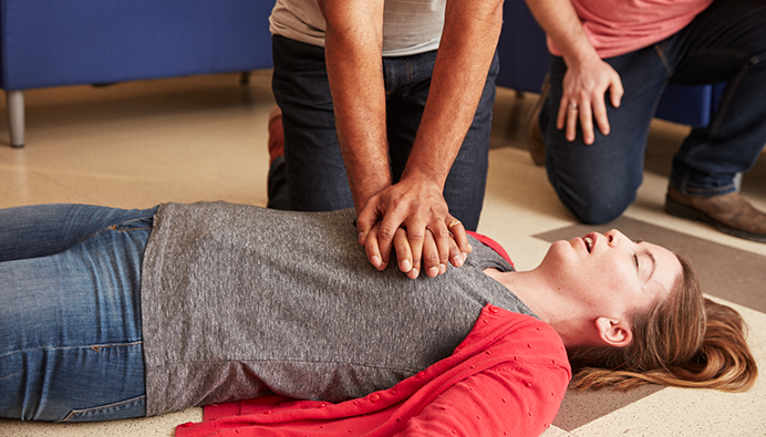 An unconscious woman lying on the floor receives CPR from a person, showing only their arms.