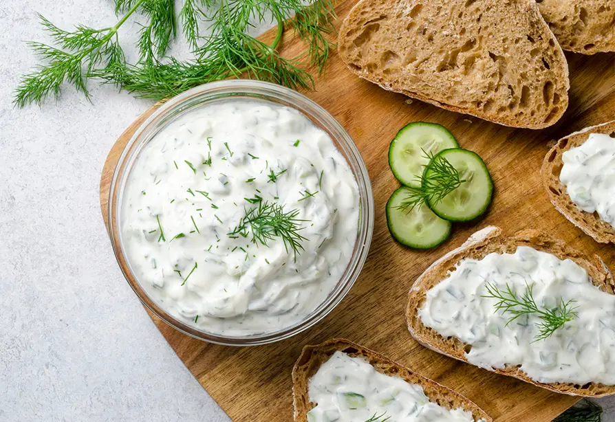 A bowl of Greek yogurt ranch dip topped with dill and spread on slices of bread on a wooden cutting board.