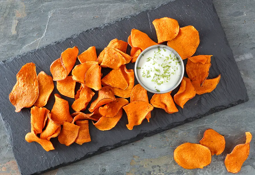 Sweet potato chips arranged on a slate platter with a bowl of yogurt dip garnished with green herbs.