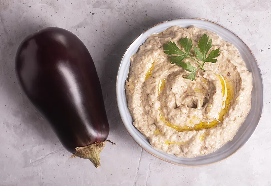 A whole eggplant next to a bowl of eggplant dip, garnished with parsley and drizzled with olive oil.