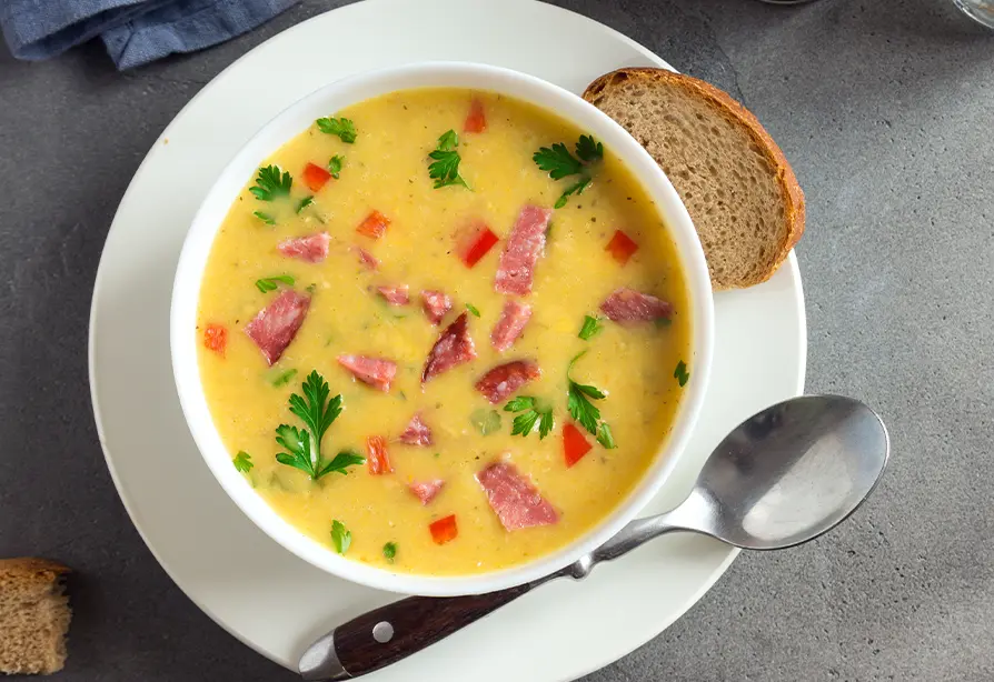 Bowl of ham and potato soup in a white bowl with a bread slice.