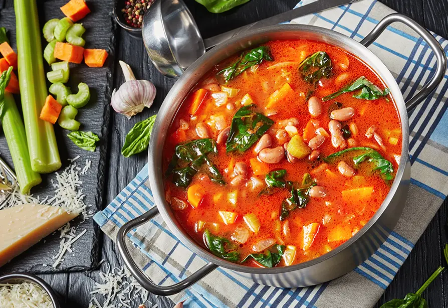 Pot of vegetable soup with beans and greens on a checkered towel.