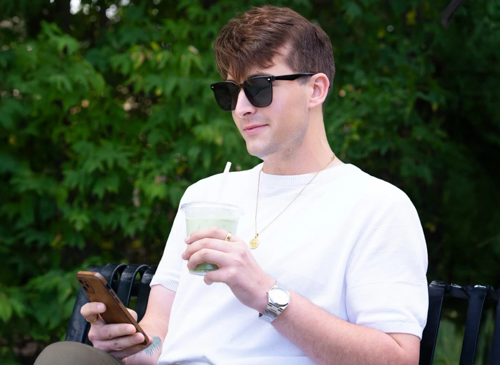 Donald browses his phone and sips juice while seated on a bench outdoors.