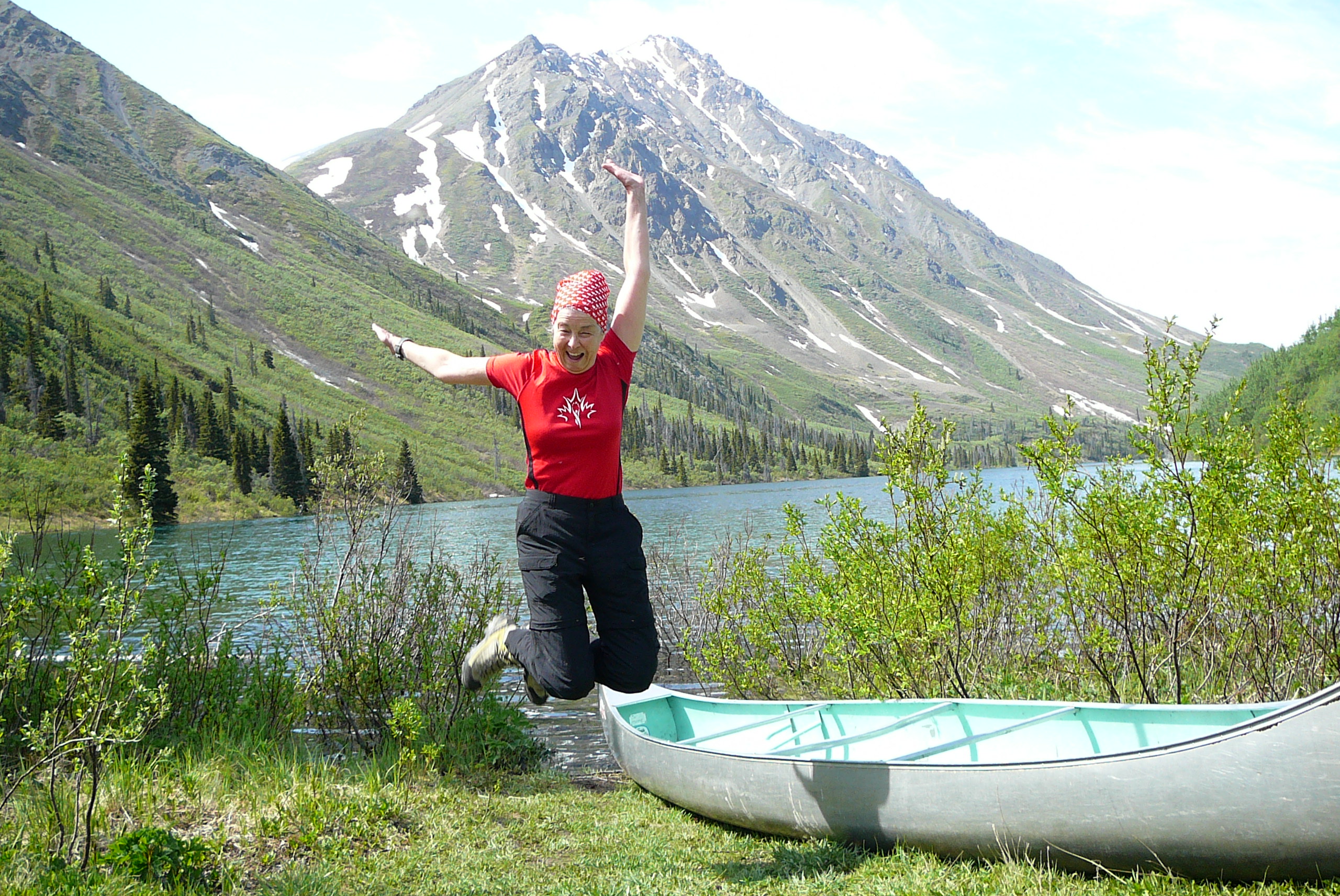 Donna smiles as she jumps in front of mountains beside a canoe. 