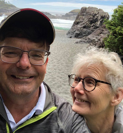 An older man and woman smile side by side on a rocky beach.