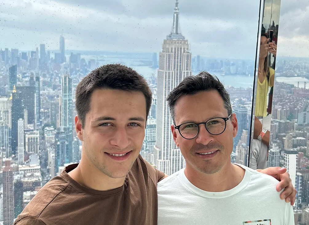 Two men stand together, arm in arm, in front of the Empire State Building.