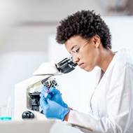 Young black woman in a lab coat looks through a microscope. 